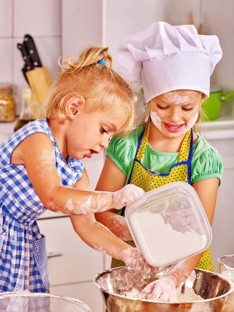 Two young girls in chef hats happily mixing dough in a kitchen.